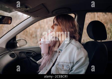 Seitenansicht der jungen modernen Frau mit rosa Frisur trinken Smoothie aus Kunststoff-Tasse mit Stroh im Auto sitzen Stockfoto