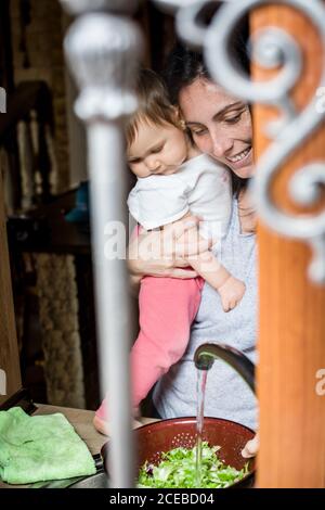 Blick durch die Fensterstangen auf lächelnde Frau, die das Baby an den Händen hält und zu Hause im Waschbecken Salat wäscht Stockfoto