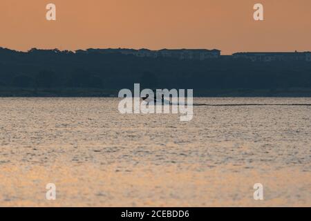 Ein Motorboot, das über einen See rast, kurz nach Sonnenaufgang mit einer hohen Eigentumswohnung, die über den Bäumen am Ufer im Hintergrund aufsteigt. Stockfoto