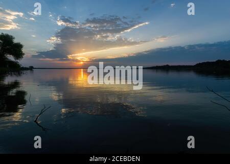 Sonnenlicht und Wolken spiegeln sich auf dem glatten See mit Bäumen an beiden Seiten bei Sonnenaufgang. Stockfoto