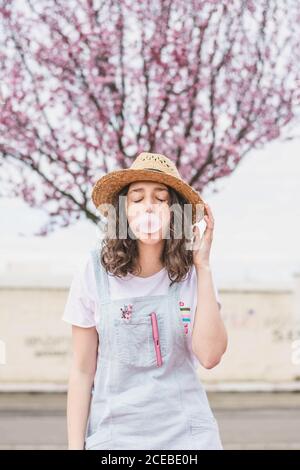 Junge romantische Teenager-Frau in Kleid und Hut machen Blase Gummi Blase gegen rosa Baum Stockfoto