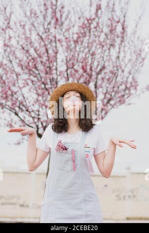 Junge romantische Teenager-Frau in Kleid und Hut machen Blase Gummi Blase gegen rosa Baum Stockfoto