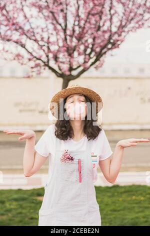 Junge romantische Teenager-Frau in Kleid und Hut machen Blase Gummi Blase gegen rosa Baum Stockfoto