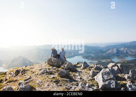 Rückansicht des jungen Paares mit kleinem Hund auf dem sitzt Spitze des rauen Felsen und Blick auf herrliche Tal mit Wunderschöne Seen an sonnigen Tagen in Spanien Stockfoto