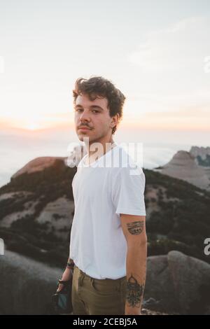 Seitenansicht eines attraktiven Typen mit Schnurrbart, der die Fotokamera hält und die Kamera anschaut, während er während des wunderschönen Sonnenaufgangs in Barcelona, Spanien, auf einer Klippe steht Stockfoto