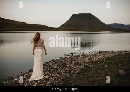 Rückansicht einer Frau in weißem Kleid, die an einem ruhigen See in Hanglage steht. Stockfoto