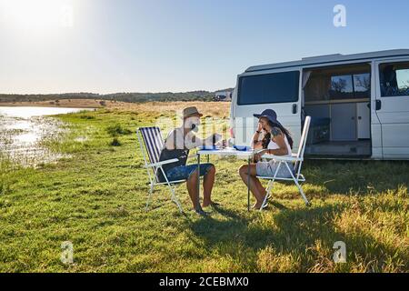 Seitenansicht Junges lächelndes Paar, das auf Klappstühlen sitzt Tisch an der Küste mit grünem Gras in der Nähe von Wasser und Automobil Im Sommer Stockfoto