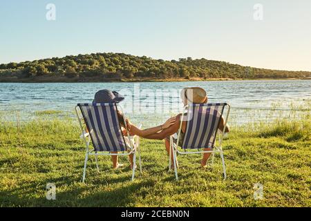 Rückansicht Junges Paar in Hüten, die Hände halten und sitzen Auf Klappstühlen an der Küste mit grünem Gras neben dem Wasser Im Sommer Stockfoto