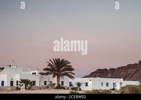 Malerischer Blick auf ein kleines weißes Hotelgebäude umgeben von Palmen im tropischen La Graciosa, Kanarische Inseln bei Sonnenuntergang mit Bergen und wunderschönem pastellfarbenen Himmel im Hintergrund Stockfoto