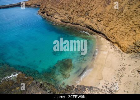 Luftaufnahme der malerischen hellen türkisfarbenen Meeresbucht, umgeben von hohen Klippen mit Wellen, die am Sandstrand am sonnigen Sommertag in La Graciosa, Kanarische Inseln, aufrollen Stockfoto
