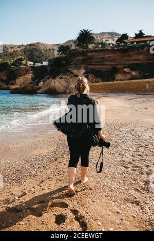 Rückansicht der jungen Barfußdame mit Fotokamera beim Gehen Auf Sand in der Nähe von Meerwasser Stockfoto