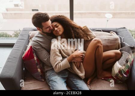 Attraktiver kaukasischer Mann, der eine hübsche afroamerikanische Frau umarmt, während er zu Hause auf einer bequemen Couch sitzt Stockfoto
