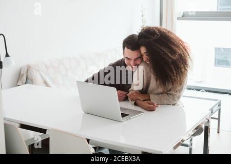 Attraktiver kaukasischer Mann und junge afroamerikanische Frau, die lächeln und zusammen auf einen modernen Laptop blicken, während sie am Tisch in einem gemütlichen Zimmer zu Hause sitzen Stockfoto