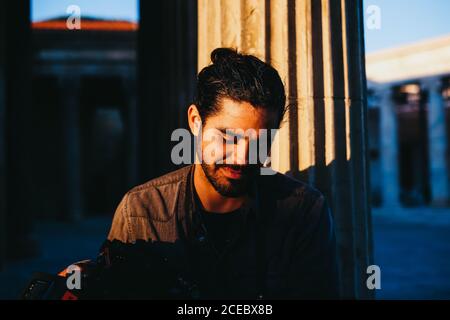 Portrait von schönen bärtigen Mann auf Stein Schritt sitzend Der Tempel im Sonnenstrahl blickt mit einem Lächeln nach unten Stockfoto