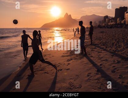 Rio de Janeiro, Brasilien - Februar 12 2014: Gruppe anonymer junger Männer, die sich gegenseitig Ball werfen, während sie sich am sandigen Ipanema-Strand in der Nähe des winkenden Meeres bei Sonnenuntergang Vergnügen Stockfoto