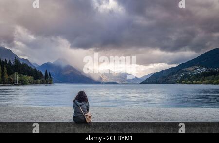 Christchurch, Neuseeland - März, 17 2015: Rückansicht der Frau in lässigem Outfit mit Blick auf ruhigen See und erstaunliche Berge am bewölkten Tag Stockfoto