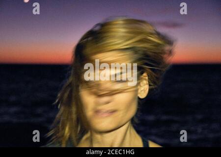 Junge Frau schüttelt blonde Haare, während sie gegen dunkle Nacht steht Himmel und wunderbares Meer Stockfoto