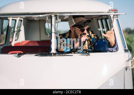 Hübscher junger Mann, der Zunge zeigt und shaka-Zeichen zeigt, während er drinnen sitzt Retro-Van auf dem Land Stockfoto