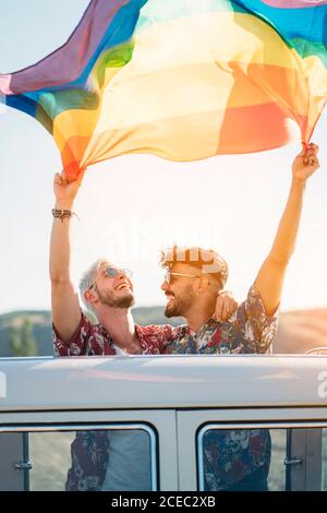 Zwei junge Männer umarmen und halten winkende LGBT-Flagge über Köpfe während?Stehen im Retro-Van mit geöffnetem Dach in der Natur Stockfoto