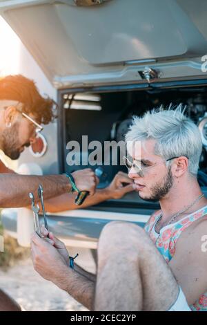 Junger Mann lehnte sich zurück und entspannen, während in der Nähe von Freund sitzen Versuchen Sie, defekten Motor des Autos während der Reise in zu reparieren Natur Stockfoto