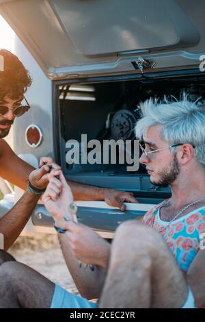Junger Mann lehnte sich zurück und entspannen, während in der Nähe von Freund sitzen Versuchen Sie, defekten Motor des Autos während der Reise in zu reparieren Natur Stockfoto