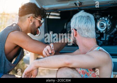 Junger Mann lehnte sich zurück und entspannen, während in der Nähe von Freund sitzen Versuchen Sie, defekten Motor des Autos während der Reise in zu reparieren Natur Stockfoto