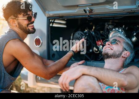 Junger Mann lehnte sich zurück und entspannen, während in der Nähe von Freund sitzen Versuchen Sie, defekten Motor des Autos während der Reise in zu reparieren Natur Stockfoto