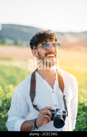 Schöner junger Kerl in Sonnenbrille fröhlich lächelnd und halten Retro Fotokamera, während Sie auf unscharfem Hintergrund der erstaunlichen Landschaft stehen Stockfoto