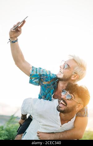 Schöner bärtiger Kerl geben Huckepack Fahrt zu fröhlichen Freund und Posiert für Selfie und verbringt Zeit in der Natur zusammen Stockfoto