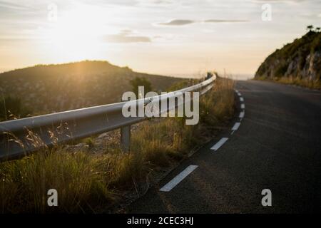 Schutzzaun und Asphaltstraße im Sonnenuntergang im Berghang beleuchtet. Stockfoto