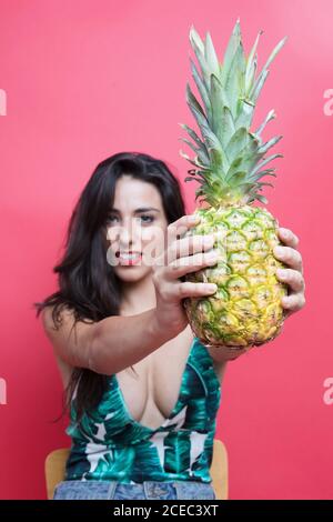 Attraktive Brünette Dame im geschnallten Hemd mit Ananas sitzend auf Stuhl und Blick auf die Kamera auf rosa Hintergrund Stockfoto
