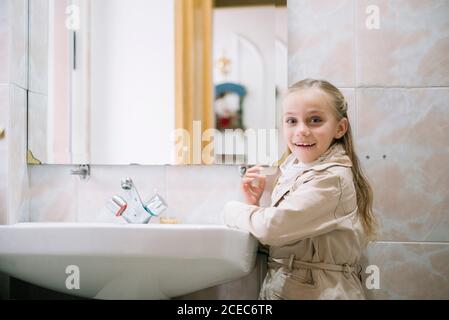 Seitenansicht des fröhlichen jungen Mädchen, das am Waschbecken im Mantel steht und die Kamera im Badezimmer anschaut. Stockfoto