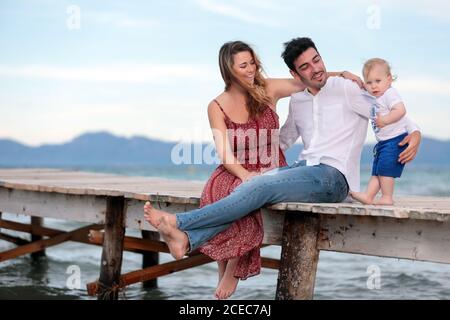 Glückliche Mutter und Vater sitzen mit kleinen Jungen auf hölzernen Pier. Stockfoto