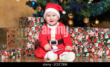 Kleiner Junge mit Kugel sitzt im Weihnachtsmann Anzug am Weihnachtsbaum. Stockfoto