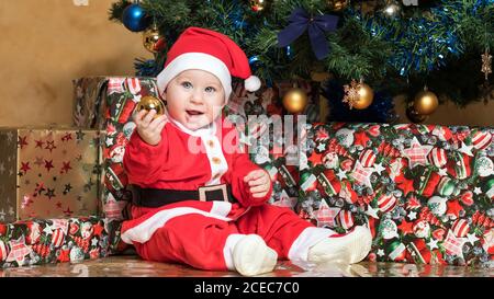 Kleiner Junge mit Kugel sitzt im Weihnachtsmann Anzug am Weihnachtsbaum. Stockfoto