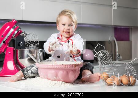 Kleiner Junge sitzt auf einem unordentlichen Tisch mit Katze und spielt mit Zutaten zum Kochen. Stockfoto