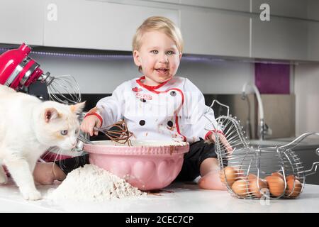 Kleiner Junge sitzt auf einem unordentlichen Tisch mit Katze und spielt mit Zutaten zum Kochen. Stockfoto