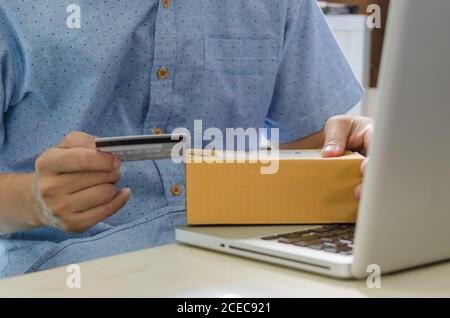 Mann Hände halten eine Kreditkarte und eine Box Paket Hämatit und Laptop am Tisch. Stockfoto