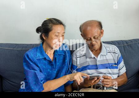 Asiatische ältere Paare beobachten Mobiltelefone und verwenden Kopfhörer auf dem Sofa. Stockfoto