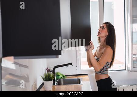 Seitenansicht einer jungen attraktiven Frau mit langen dunklen Haaren, die in der Küche steht und in einen schwarzen, geöffneten Schrank schaut Stockfoto