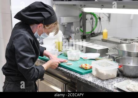 Frau Chef in Schutzmaske Gesicht bereiten Essen in der Küche eines Restaurants oder Hotels. Coronavirus-Präventionskonzept. Hochwertige Fotos Stockfoto