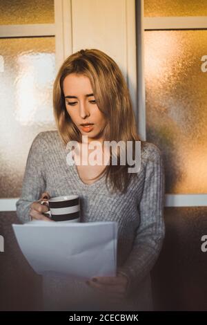 Schockierte junge Frau, die zu Hause einen Becher stand und Papiere las. Stockfoto
