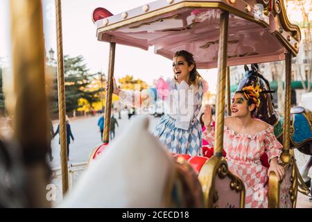 Zwei hübsche junge Frauen mit Theaterschminke und Kostümen lachen Beim Sitzen auf erstaunlichen Kreisverkehr im Vergnügungspark Stockfoto