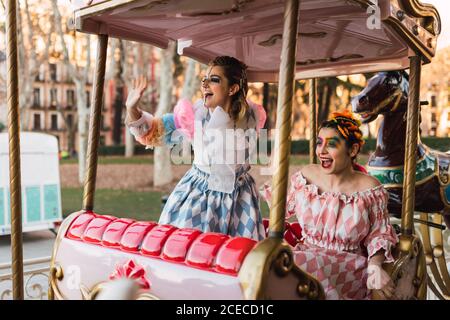 Zwei hübsche junge Frauen mit Theaterschminke und Kostümen lachen Beim Sitzen auf erstaunlichen Kreisverkehr im Vergnügungspark Stockfoto