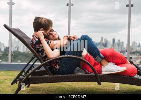 Seitenansicht von niedlichen jungen Paar Augen geschlossen halten Und kuscheln, während Sie auf einer bequemen Liege auf der Terrasse liegen Moderne Stadt Stockfoto