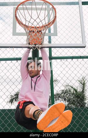 Von unten Aufnahme von fröhlichen jungen Kerl Blick auf die Kamera Beim Aufhängen am Basketballring, während Sie Zeit auf Sport verbringen Masse Stockfoto