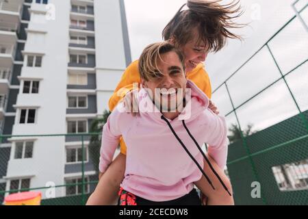 Schöner junger Kerl lachend und geben Huckepack Fahrt aufgeregt Freundin, während Sie Zeit auf dem Sportplatz zusammen verbringen Stockfoto