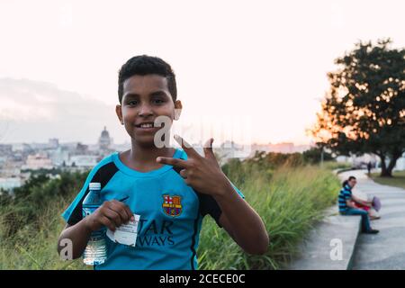 LA HABANA, KUBA - 6. NOVEMBER , 2018: Hispanic Junge in Fußball-Trikot mit Flasche Wasser in der Nähe Teenager zeigt zwei und Blick auf Kamera in Kuba Stockfoto