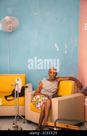 LA HABANA, KUBA - 6. NOVEMBER 2018: Älterer afroamerikanischer Mann sitzt auf einem Sessel in der Nähe von Spazierstöcken im Raum in der Nähe der blauen Wand in Kuba Stockfoto