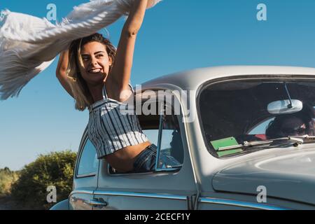 Fröhliche junge Frau, die aus dem Autofenster ragt und einen winkenden leichten Schal hält, der die Reise in hellem Sonnenlicht genießt Stockfoto
